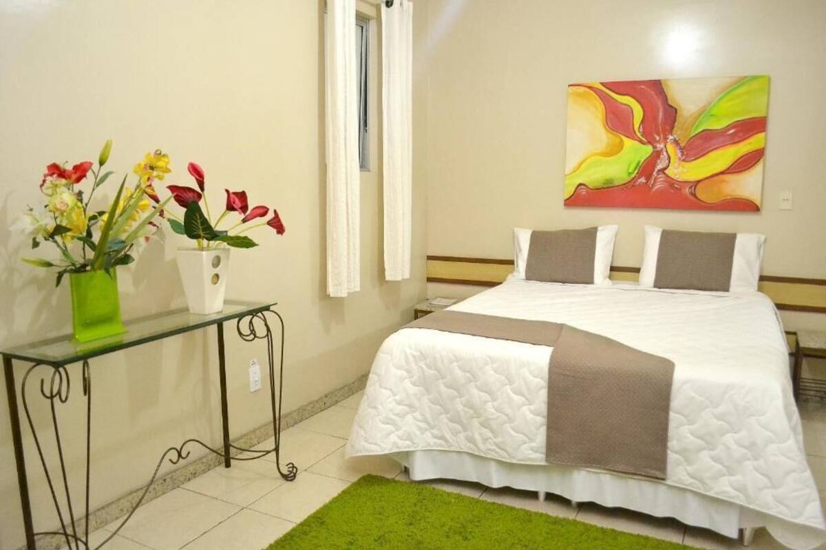 A hotel room with a bed, a glass table with flowers, a painting on the wall, and a green rug.