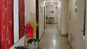 A hallway with a red wall, a door, a plant with red and yellow flowers, and a dining area in the background. - Domus Hotel Veneza (Ipatinga)