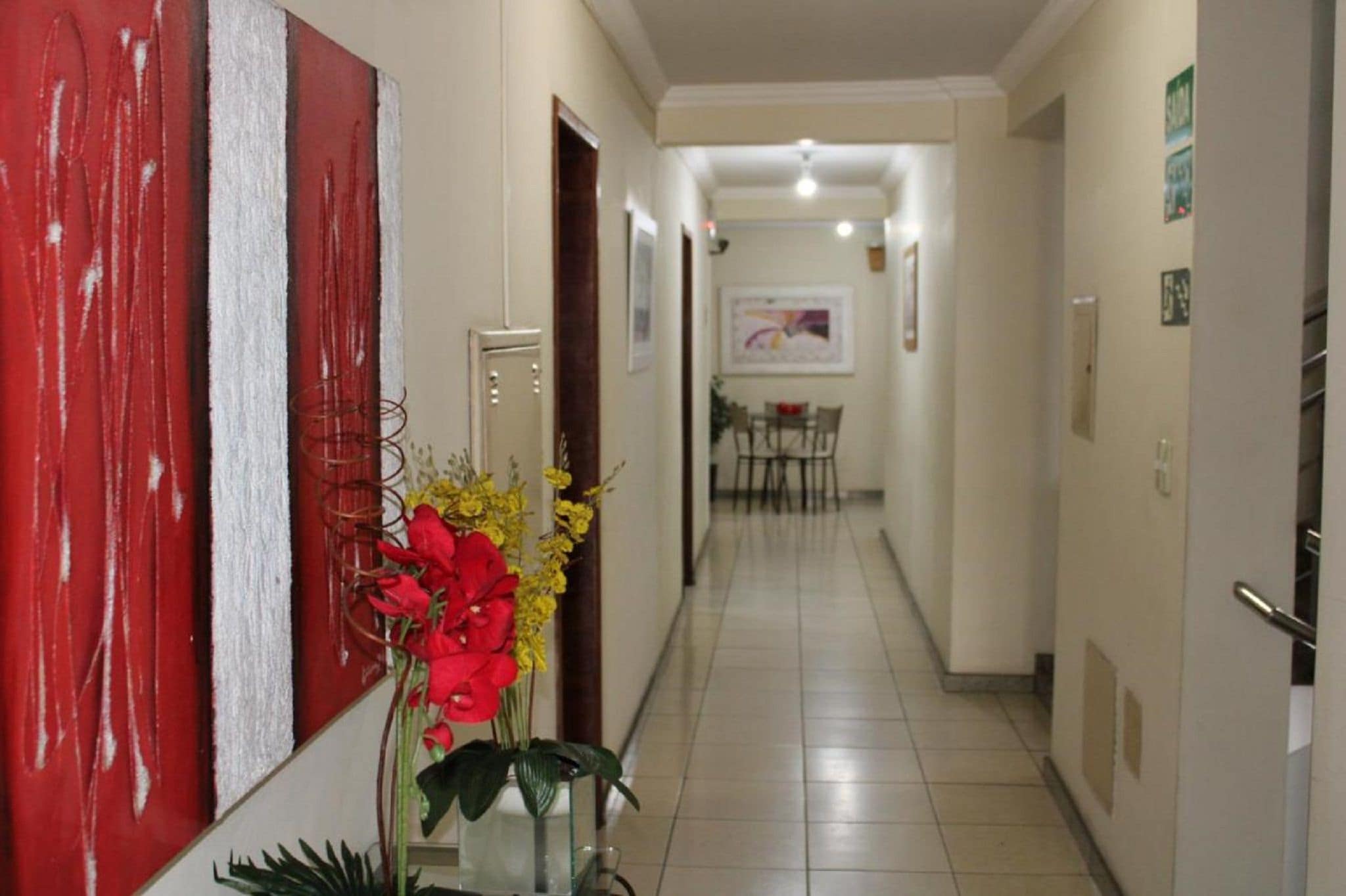 A hallway with a red wall, a door, a plant with red and yellow flowers, and a dining area in the background.