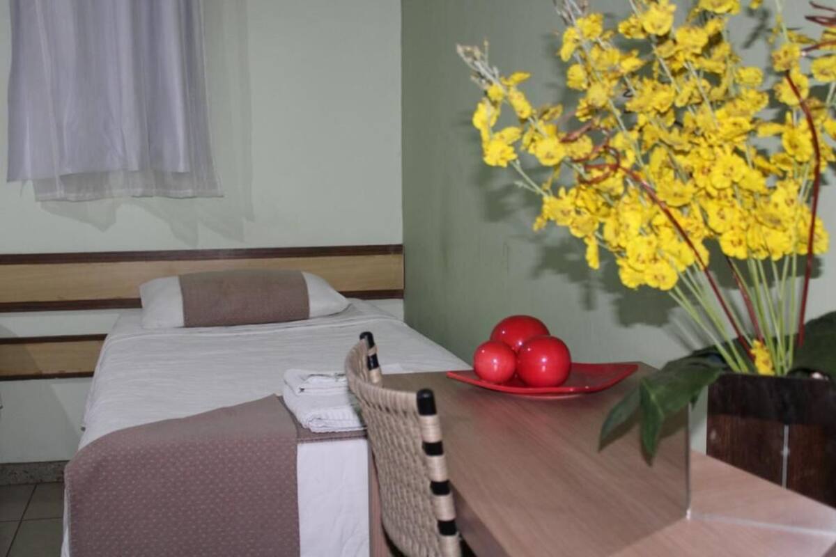 A hotel room with a bed, a wooden nightstand, a chair, a red tray with red balls, and a yellow flower arrangement.