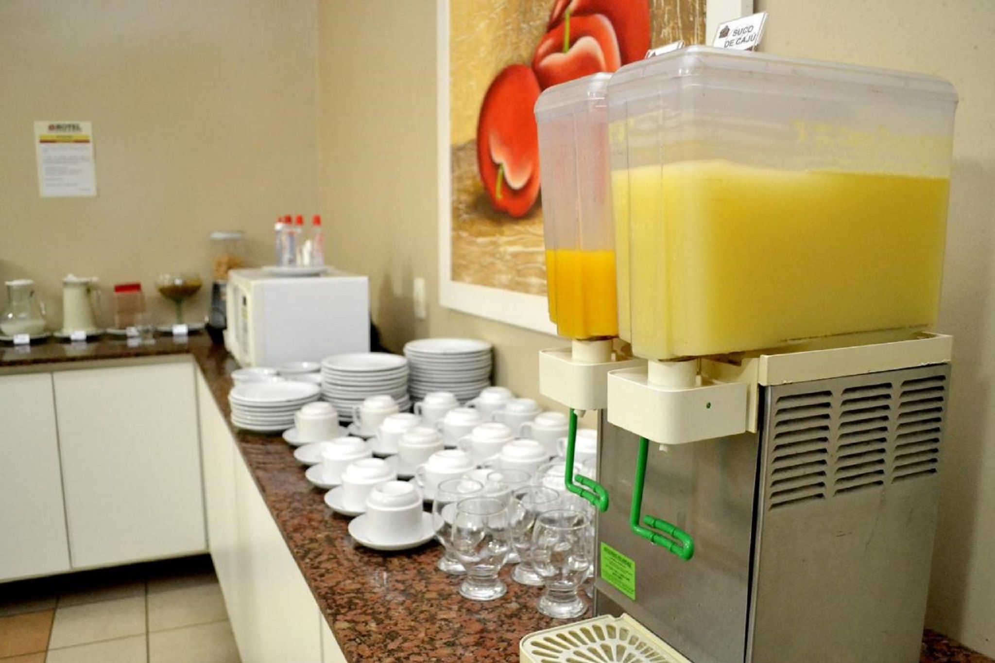 Um dispensador de suco com um recipiente grande, uma geladeira e um balcão com pratos e copos empilhados.
