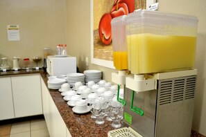 Un distributeur de jus avec un grand réservoir, un réfrigérateur et un comptoir où sont empilées des assiettes et des verres.