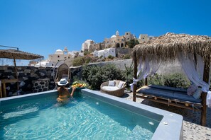 A pool area with a stone wall, a wicker chair, a cushioned lounge, and a thatched umbrella. - Rock Villa Legend: Modern Comfort in Timeless Caves /3BR  (Thira - Santorini)
