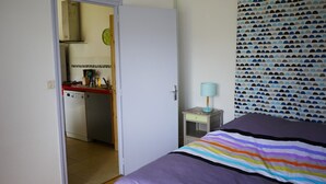 Ein Schlafzimmer mit einem Bett, einem Nachttisch mit Lampe, einem gemusterten Vorhang und Blick zur Küche.