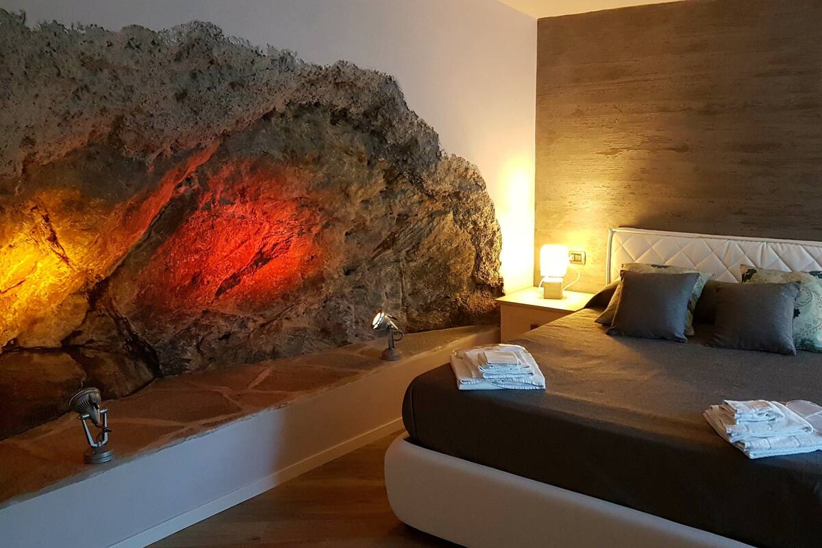 A bedroom with a large rock wall, a white headboard, and a bedside table with a lamp.