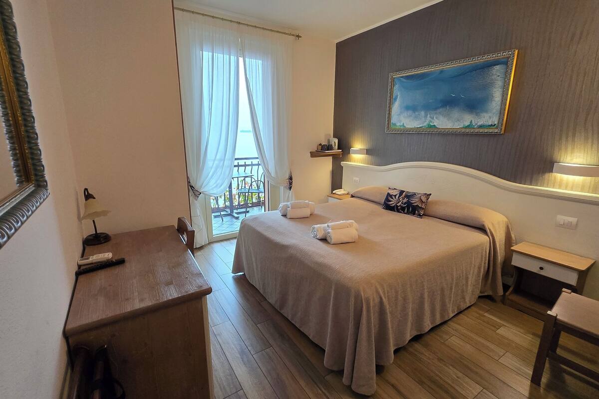 A hotel room with a bed, bedside tables, a desk, a chair, a painting, and a view of the outdoors.
