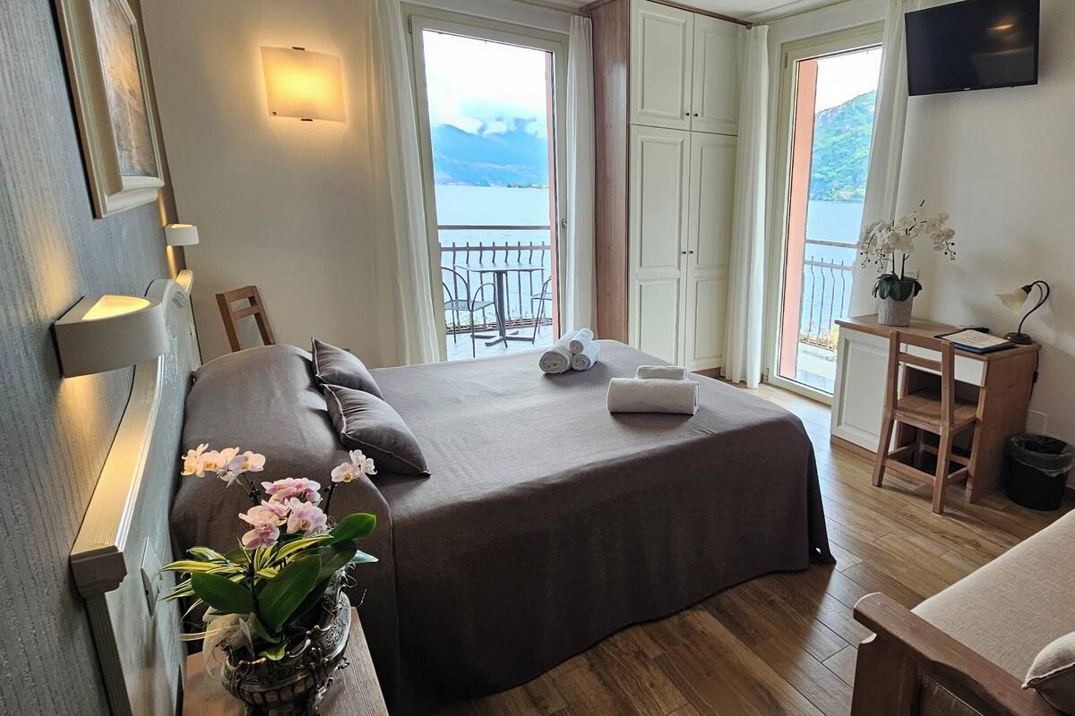A bedroom with a bed, a small desk, a chair, a sofa, a potted orchid, and a view of the outdoors.