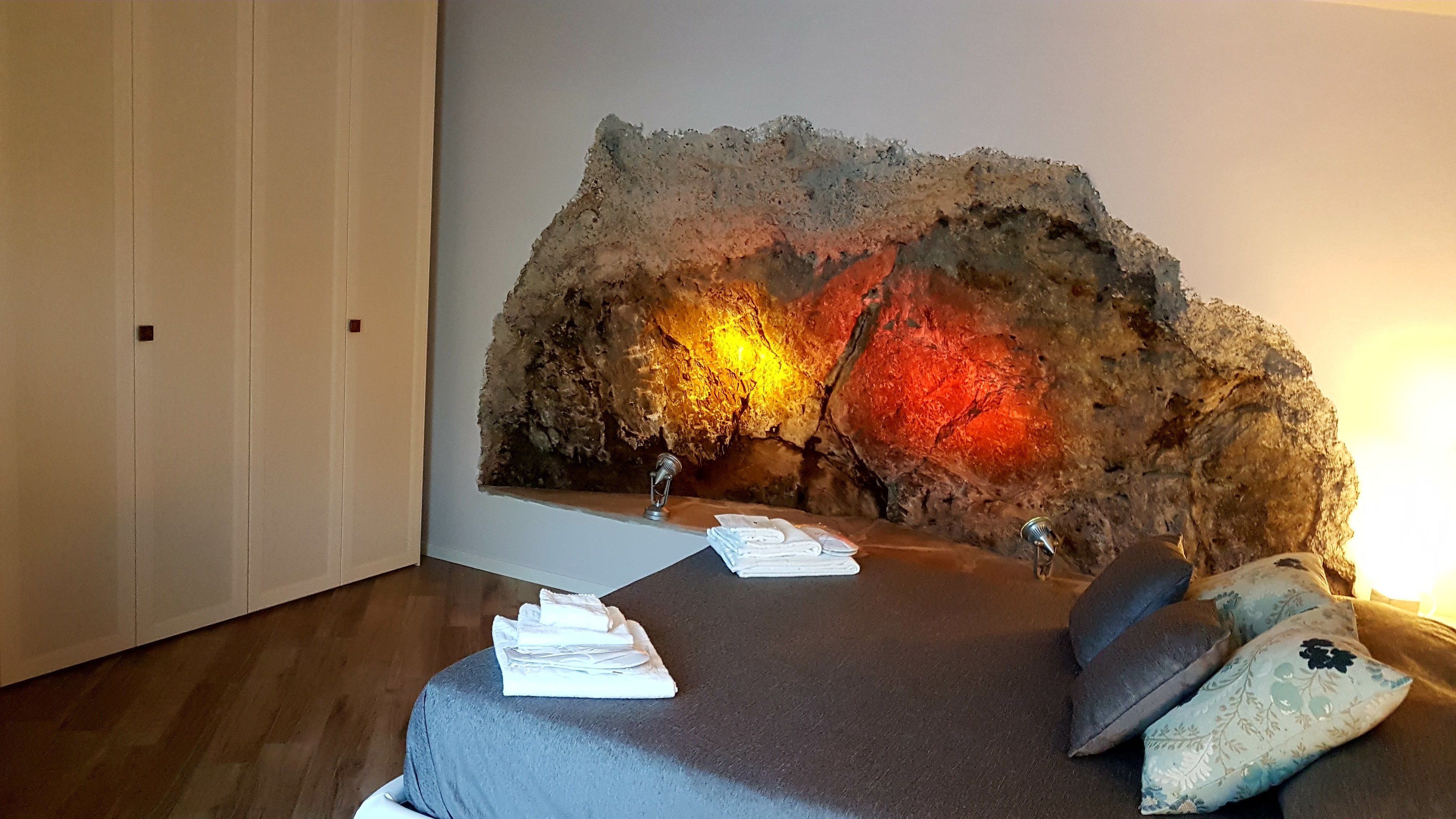 Quarto com uma cama grande, parede de pedra iluminada e uma pequena mesa com toalhas.