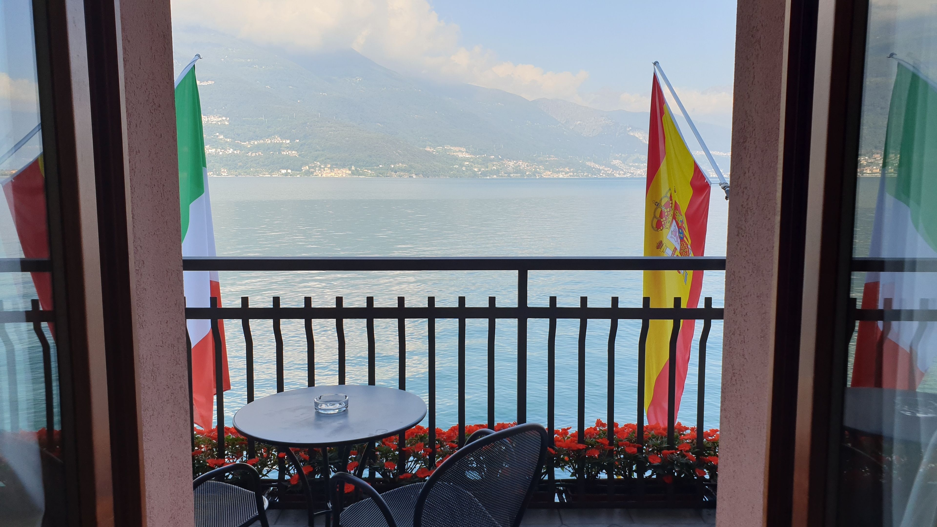 Un balcon avec une table et des chaises, d’où l’on peut admirer un plan d’eau et des montagnes. On peut voir des drapeaux sur le balcon.
