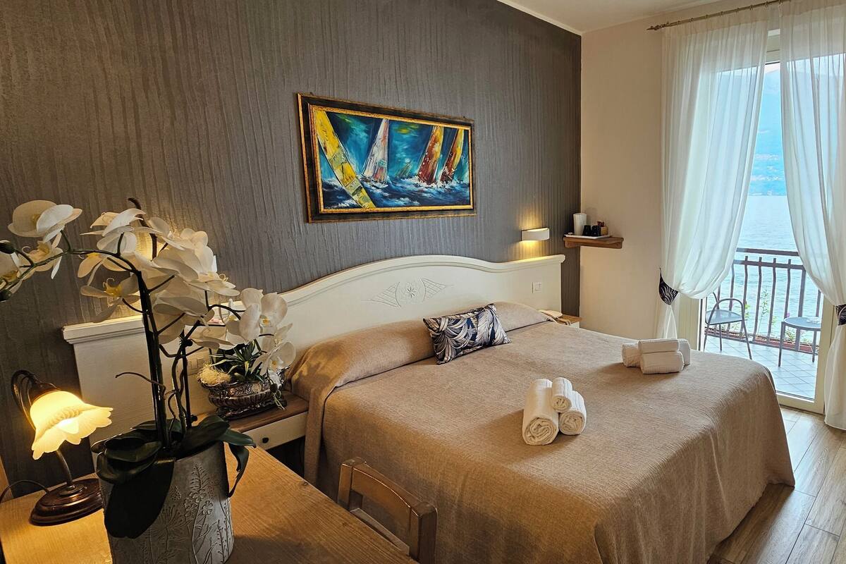 A hotel room with a bed, a nightstand, a lamp, a painting, and a view of the sea through the window.