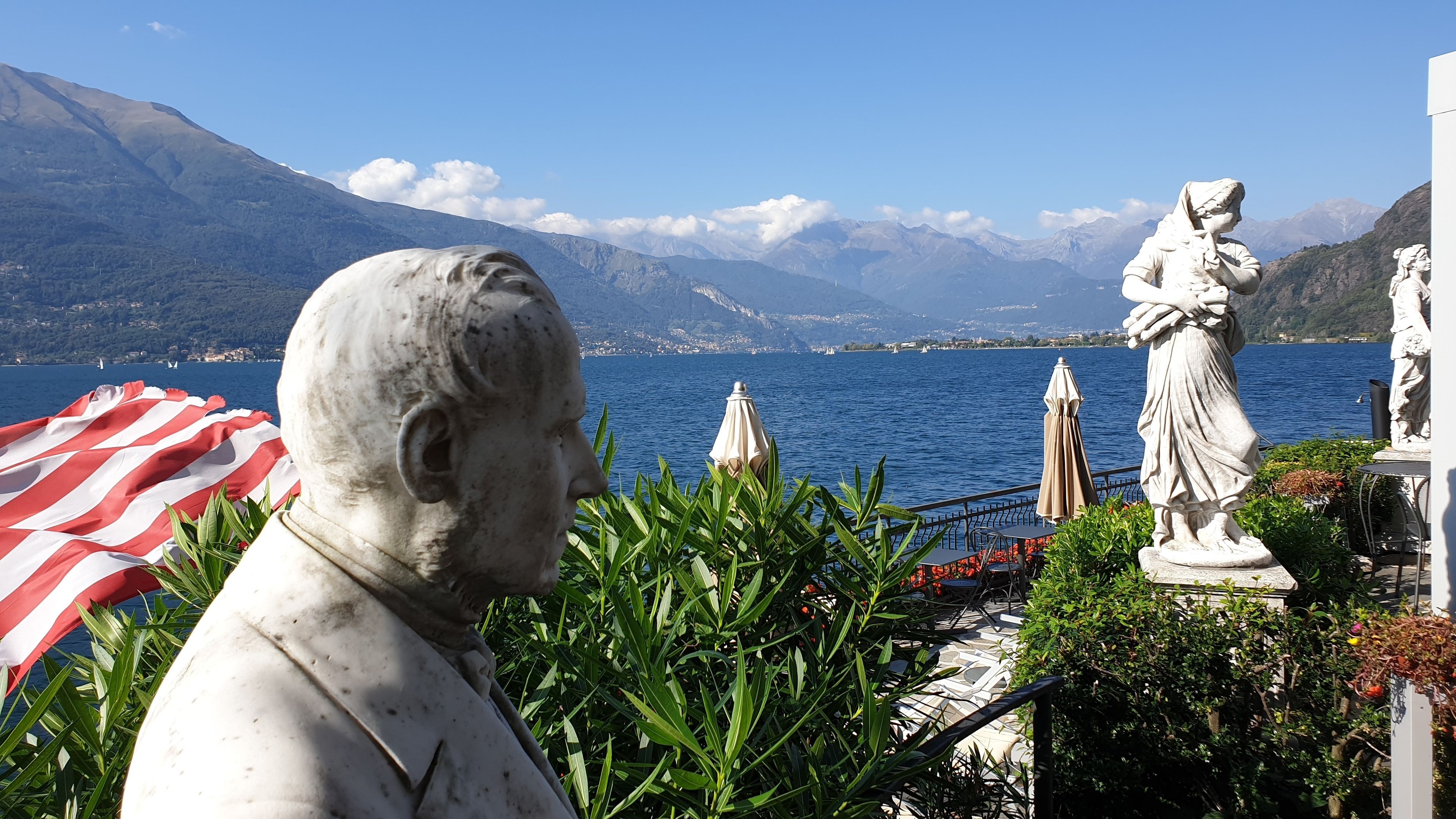 A statue of a man in a white suit, a red and white striped umbrella, and a view of a lake with mountains in the background.