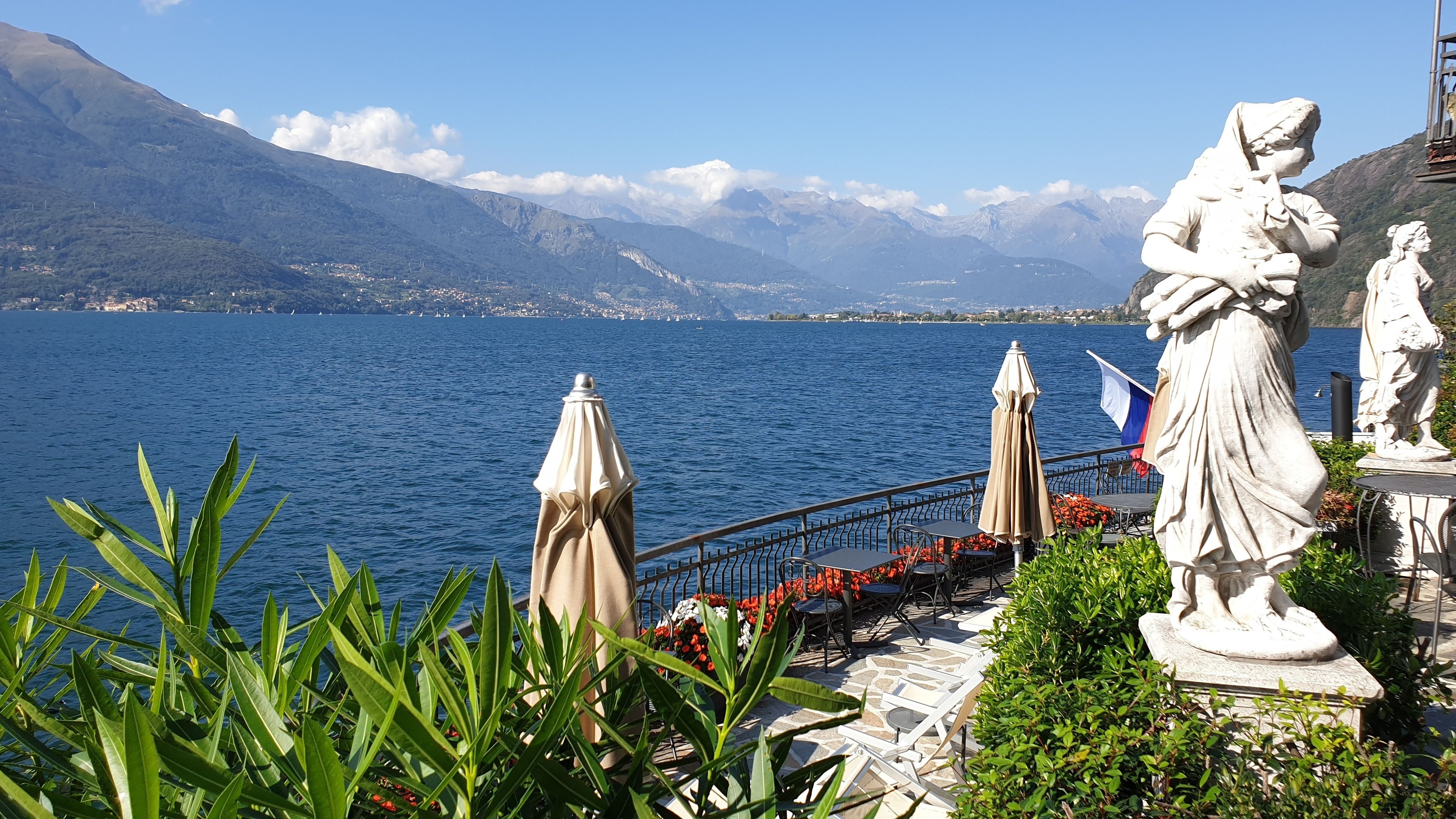 Terraza junto al lago con mobiliario exterior, una estatua y vistas a montañas y un pueblo distante.