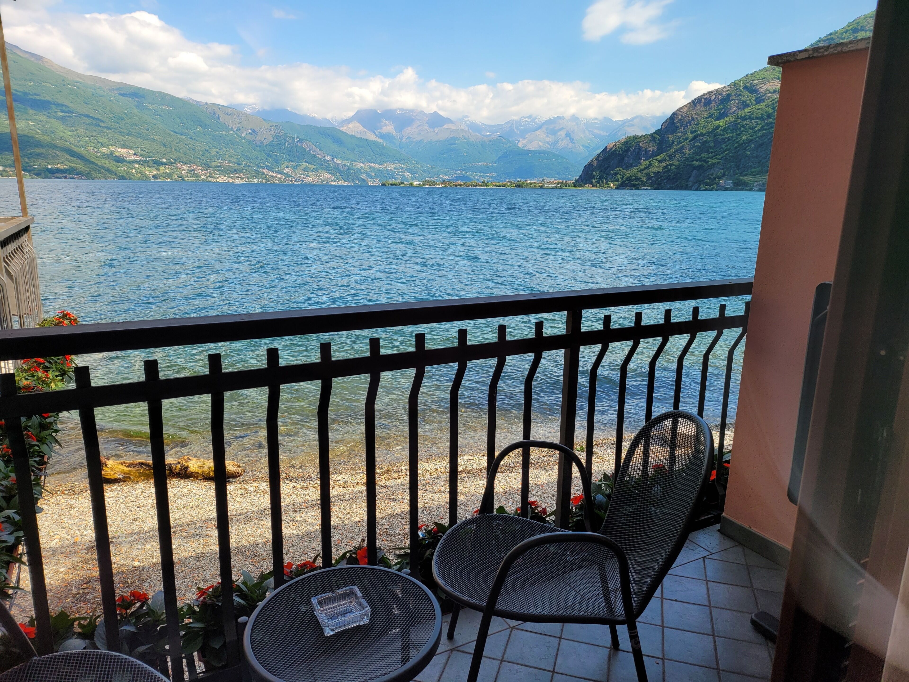 A balcony with a view of a lake and mountains. There are two chairs and a table on the balcony.