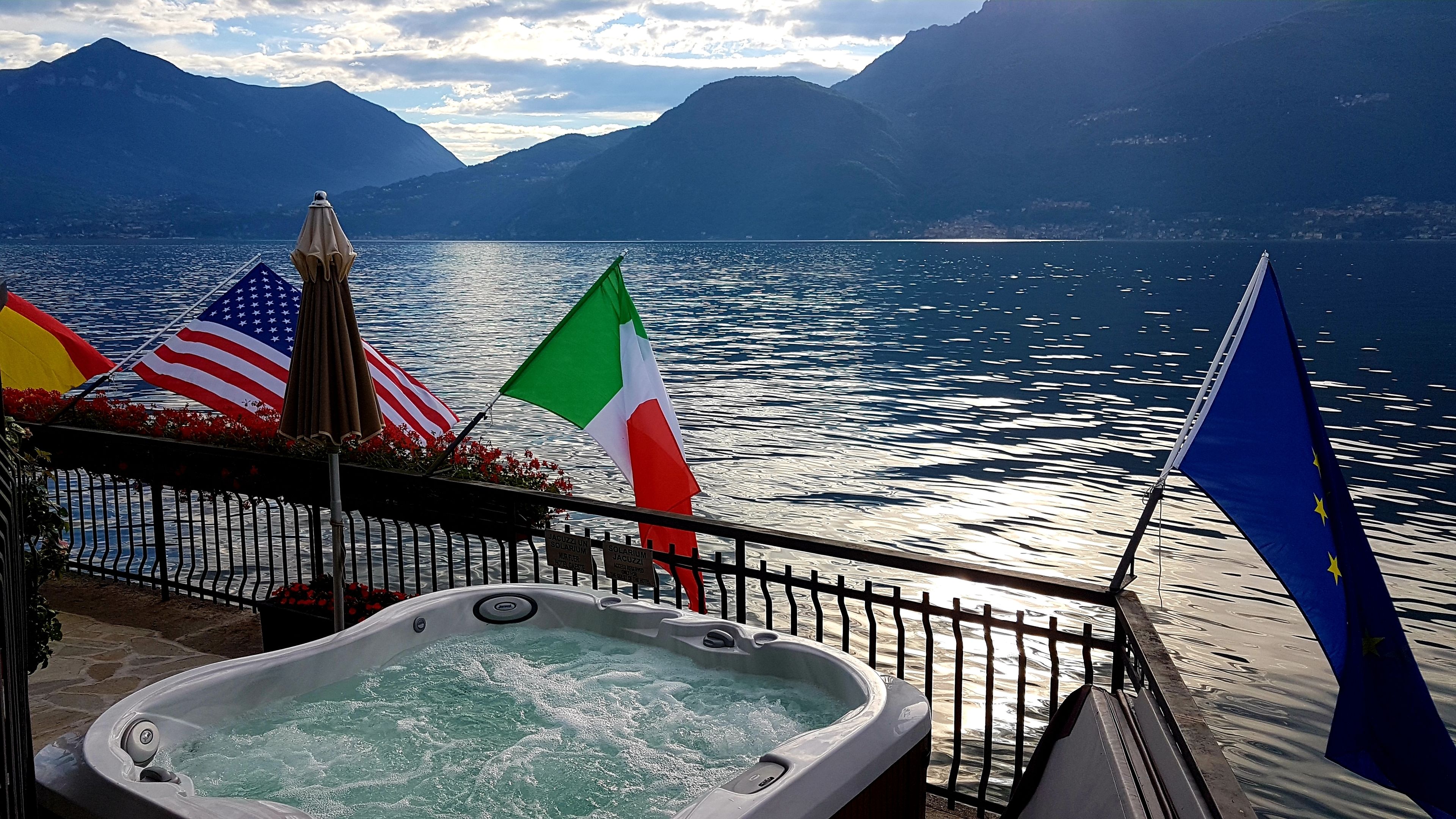 Ein Whirlpool mit Blick auf einen See und Berge, Flaggen der USA, Italiens und der EU, sowie ein Schild mit der Aufschrift 38°C Wassertemperatur.