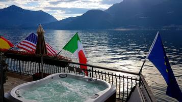 Ein Whirlpool mit Blick auf einen See und Berge, Flaggen der USA, Italiens und der EU, sowie ein Schild mit der Aufschrift 38°C Wassertemperatur.
