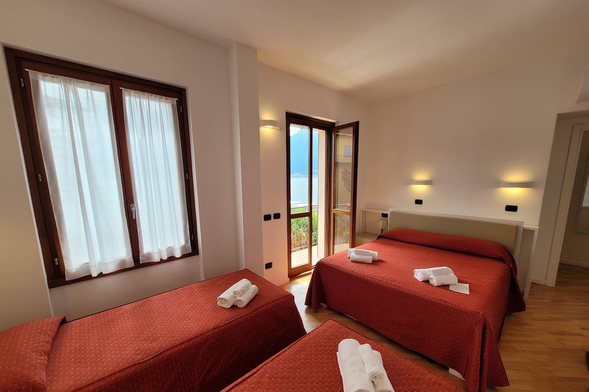 A hotel room with two single beds, each with a red bedspread and white towels. There is a balcony with a glass door leading outside.