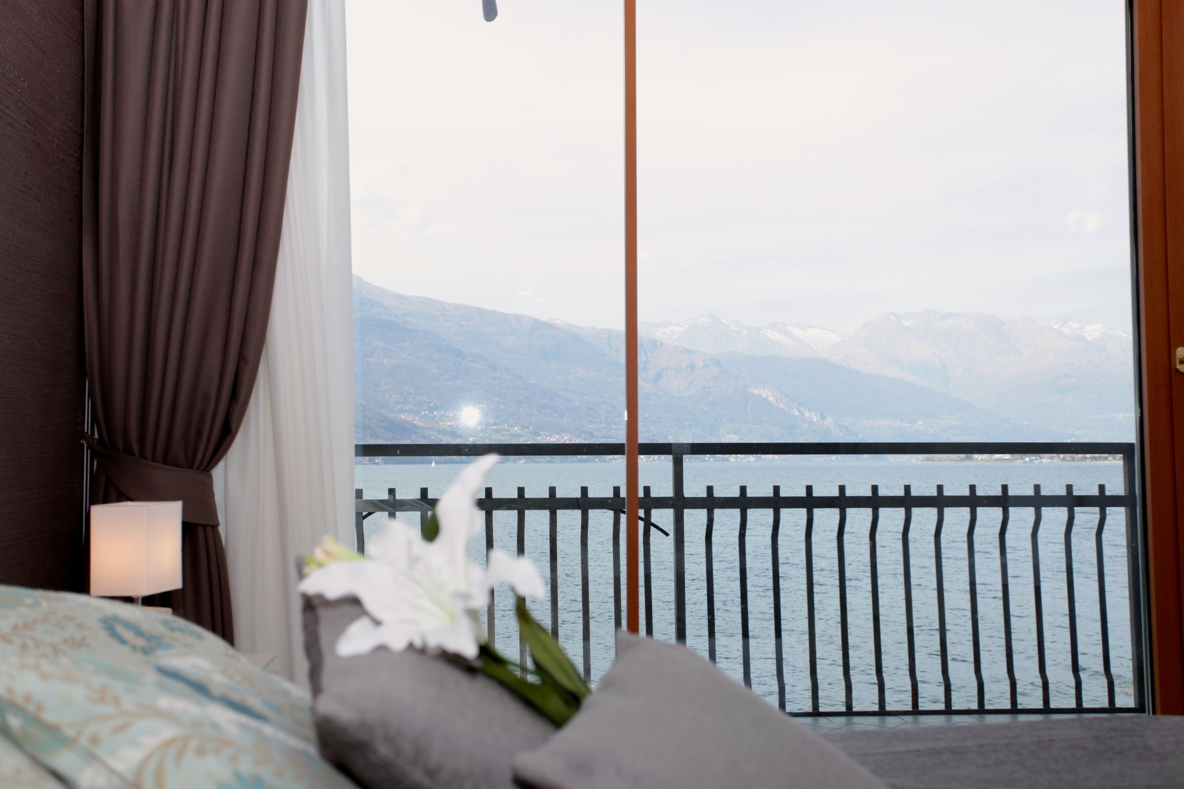 A hotel room with a bed, a nightstand, a lamp, a flower arrangement, and a view of a lake with mountains in the background.