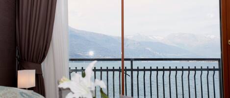A hotel room with a bed, a nightstand, a lamp, a flower arrangement, and a view of a lake with mountains in the background.