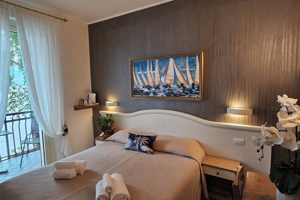 A hotel room with a bed, a painting of sailboats, a balcony with a view, and a vase of orchids.