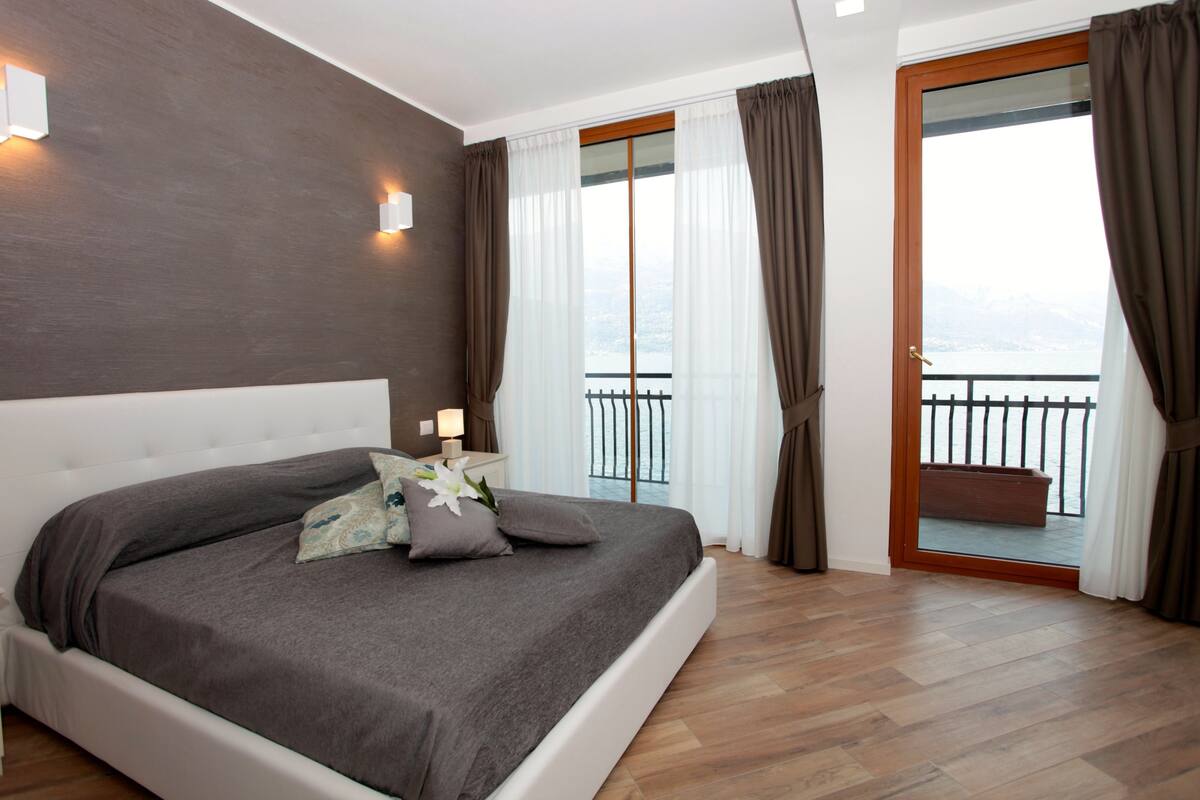 A bedroom with a large bed, a nightstand, a window with curtains, and a balcony with a railing.