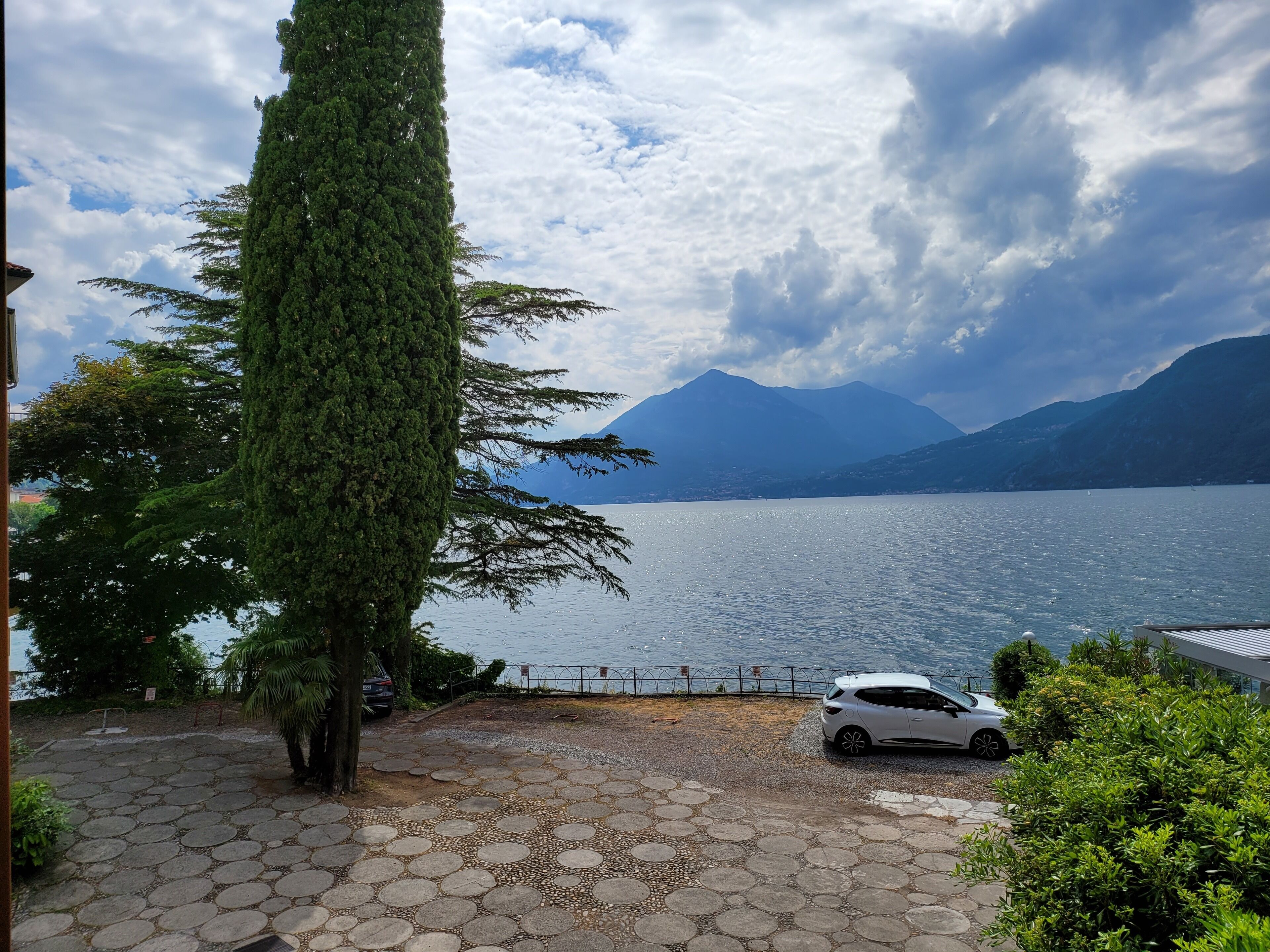 Une zone pavée où est garée une voiture, un grand arbre et une vue sur un lac avec des montagnes en arrière-plan.