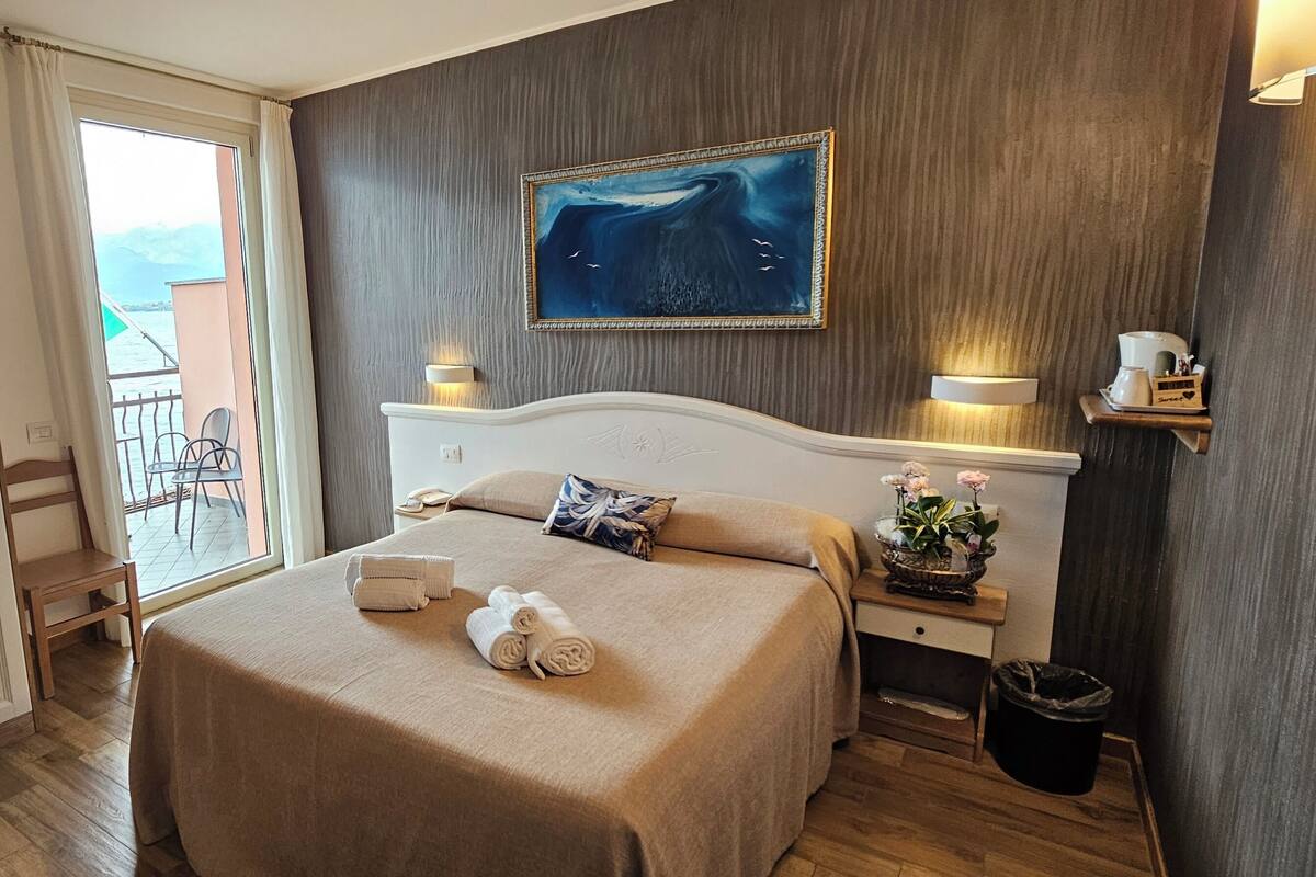 A hotel room with a bed, a painting, a nightstand, a chair, and a view of the outdoors.