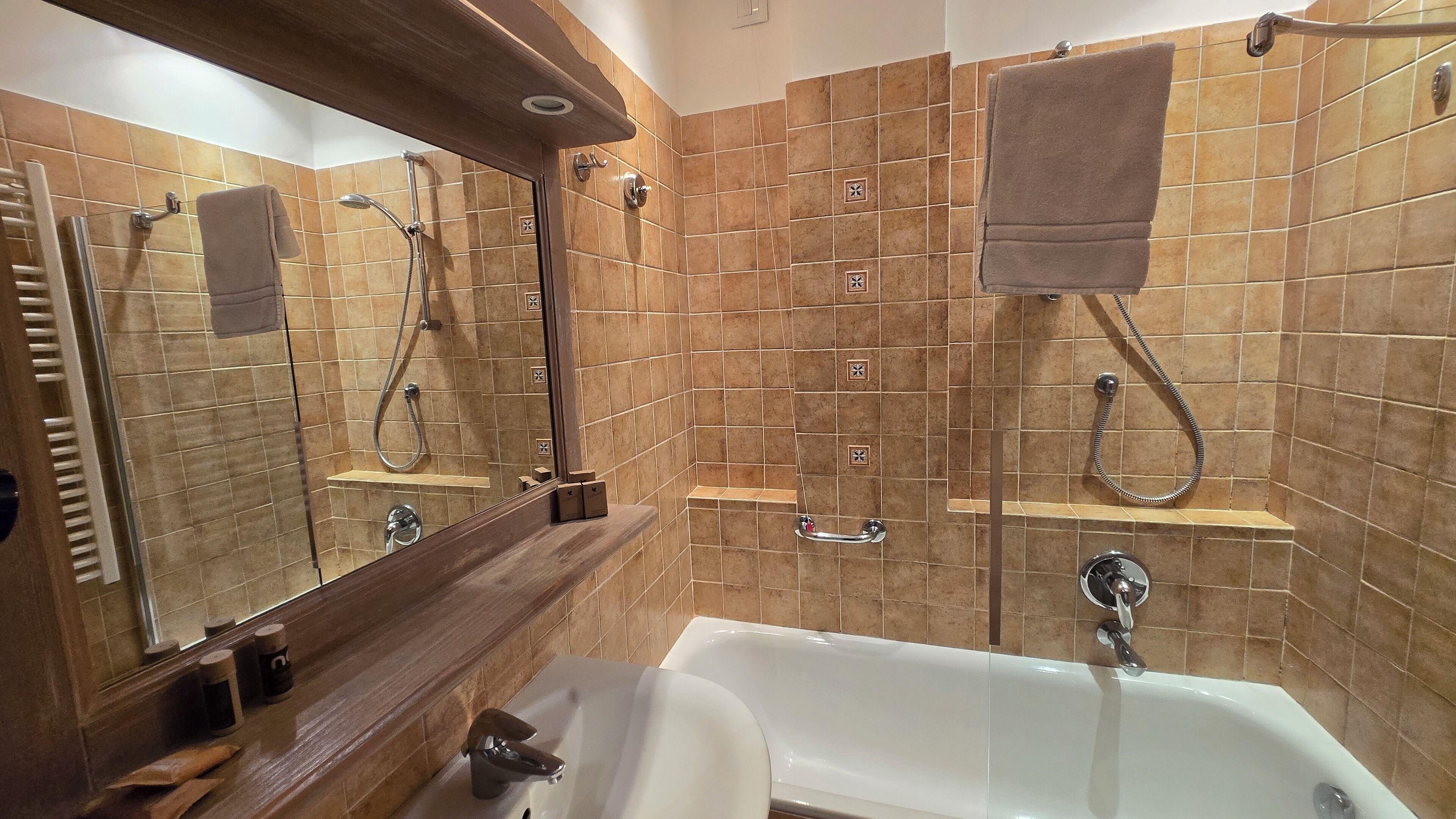Ein Badezimmer mit einer Badewanne, zwei Duschen, einem Waschtisch mit Spiegel und braunen Fliesenwänden.