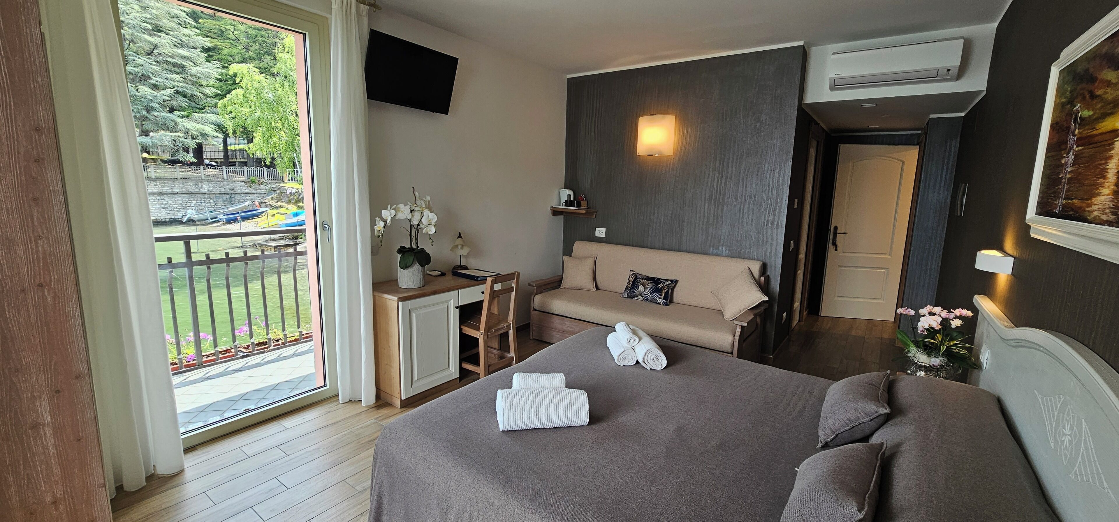 Een hotelkamer met een bed, een slaapbank, een bureau met een lamp, een televisie en een balkon met uitzicht op de omgeving.