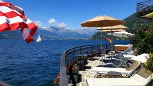 A boat deck with lounge chairs, umbrellas, and a view of a sailboat on a lake with mountains in the background. - Meridiana Boutique Hotel (Bellano)