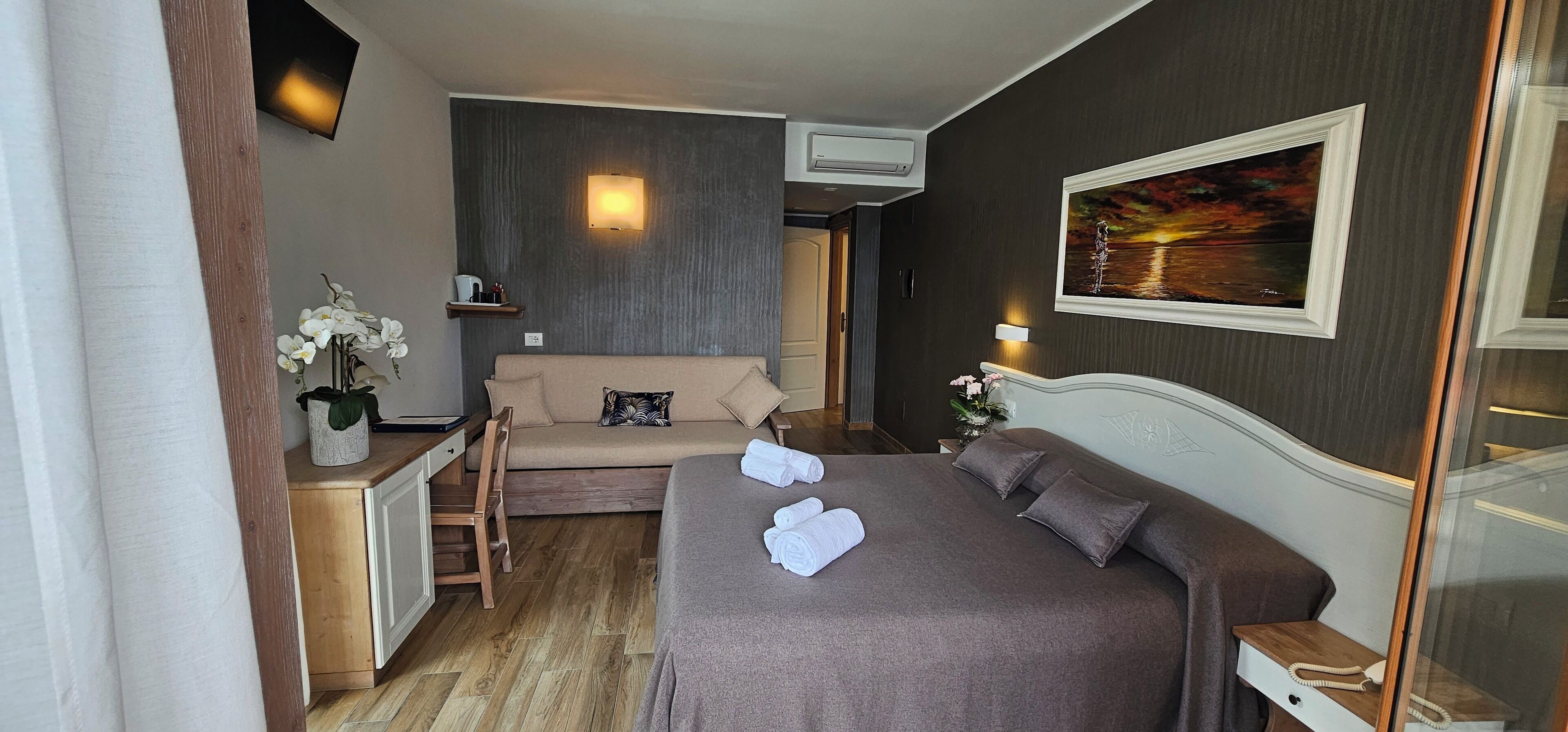 A hotel room with a bed, a sofa, a desk with a chair, a vase with flowers, and a painting on the wall.