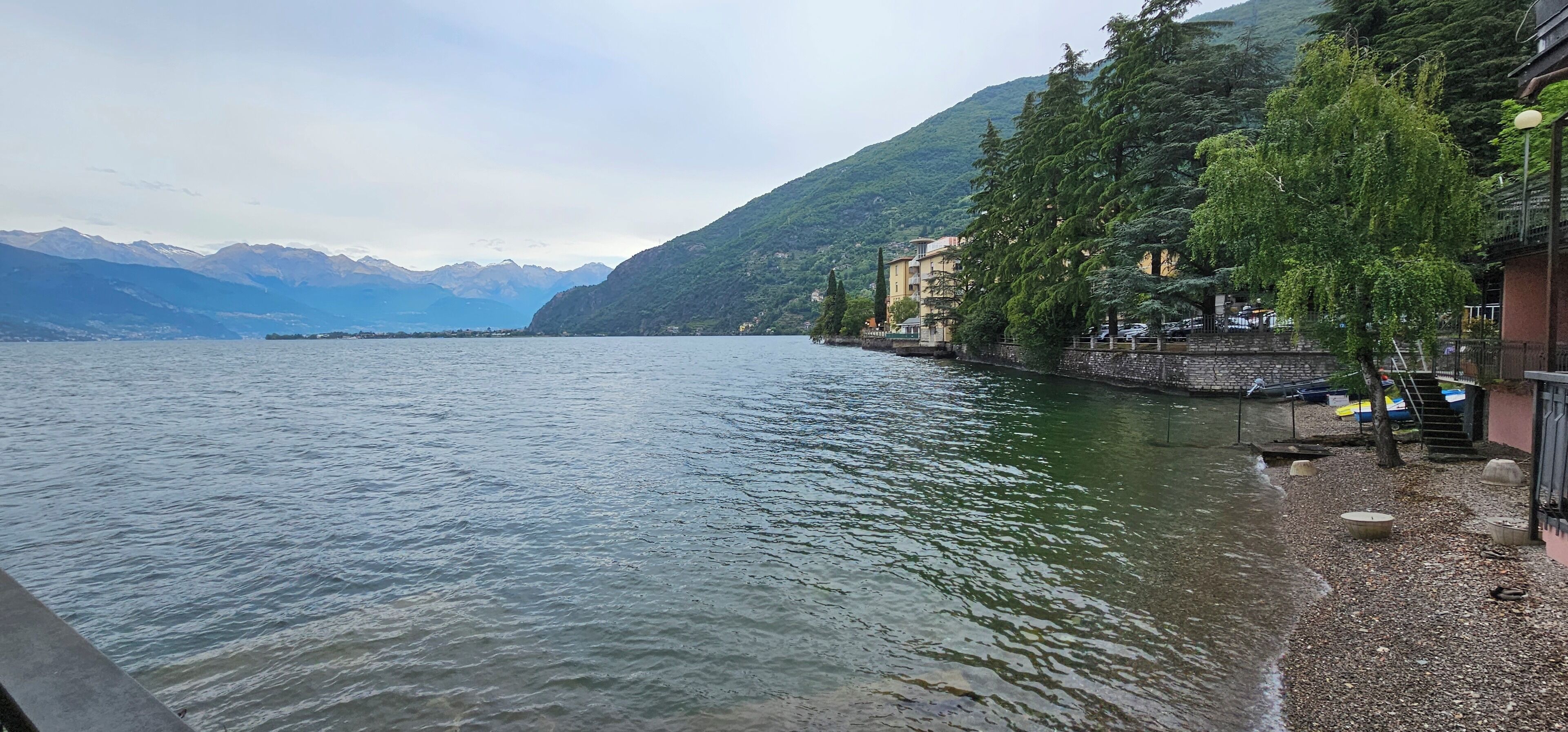 A lakeside view with buildings along the shore, clear water, and mountains in the background.