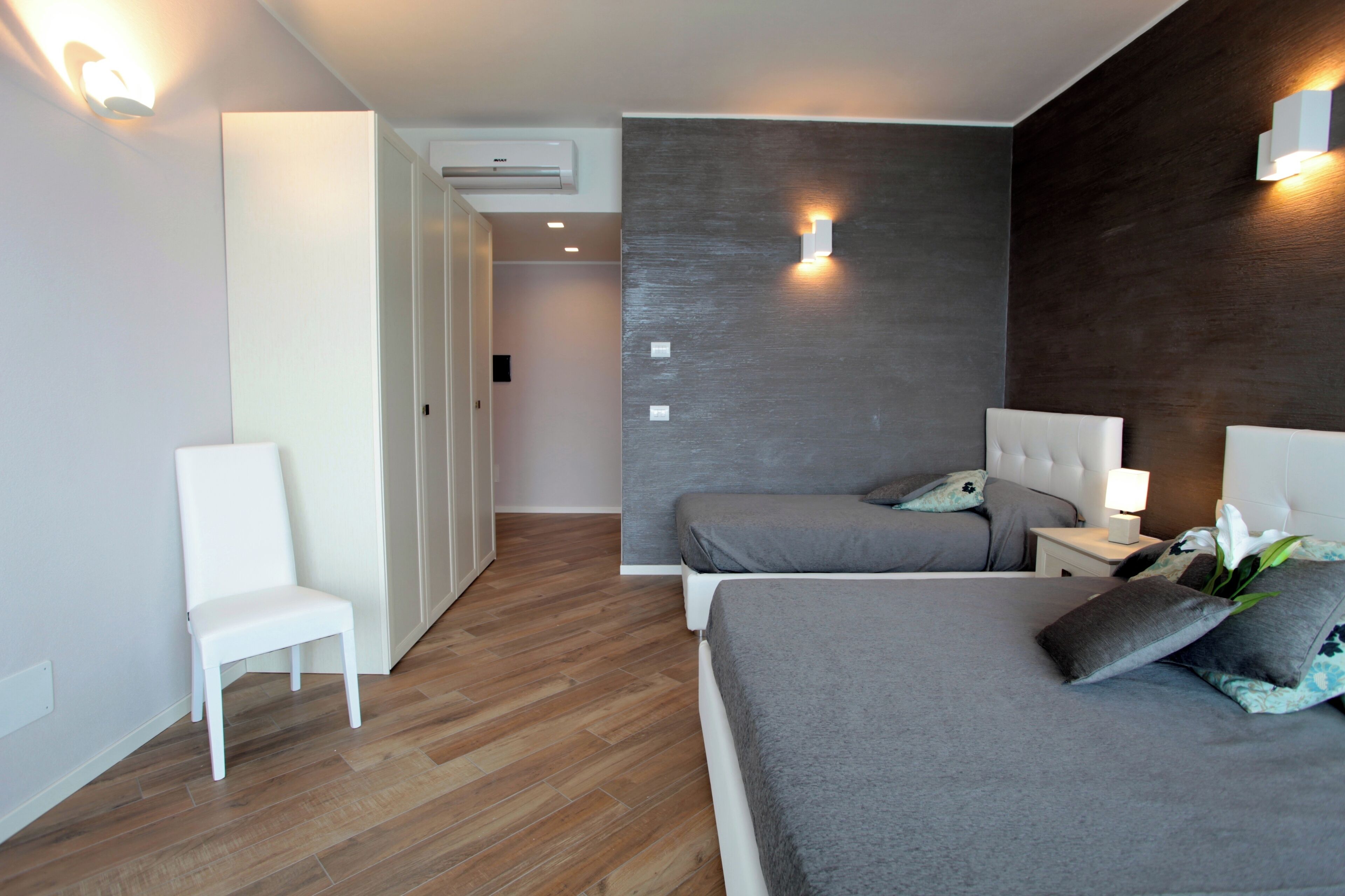 A modern hotel room with a wooden floor, a white chair, two beds with grey bedding, and a dark wooden accent wall.