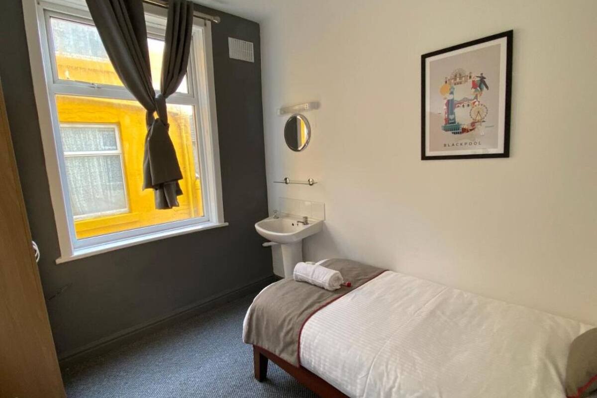 A small, clean room with a single bed, a bedside table, a window with curtains, a sink, and a framed picture on the wall.