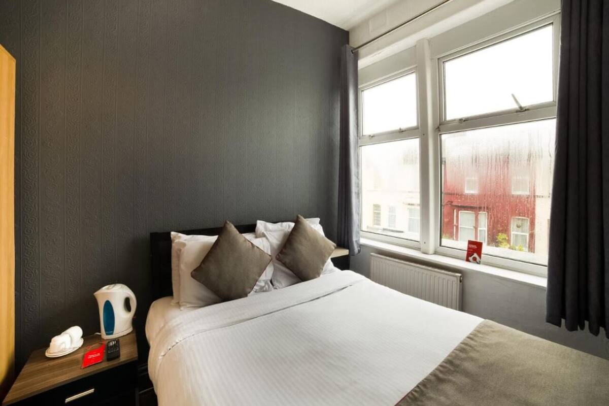 A hotel room with a large bed, a nightstand with a kettle, a telephone, and a window with a view of buildings.