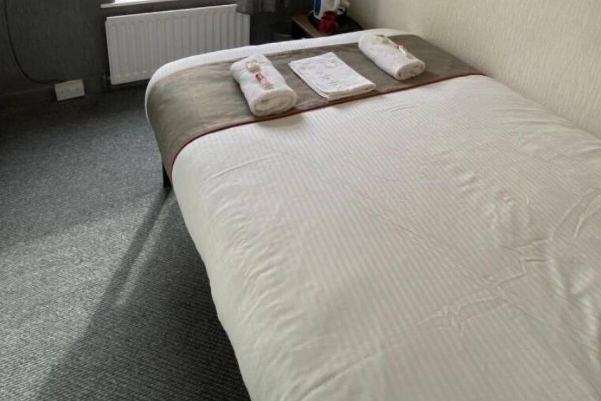 A hotel room with a bed, a window with curtains, a radiator, and a small table with a kettle.