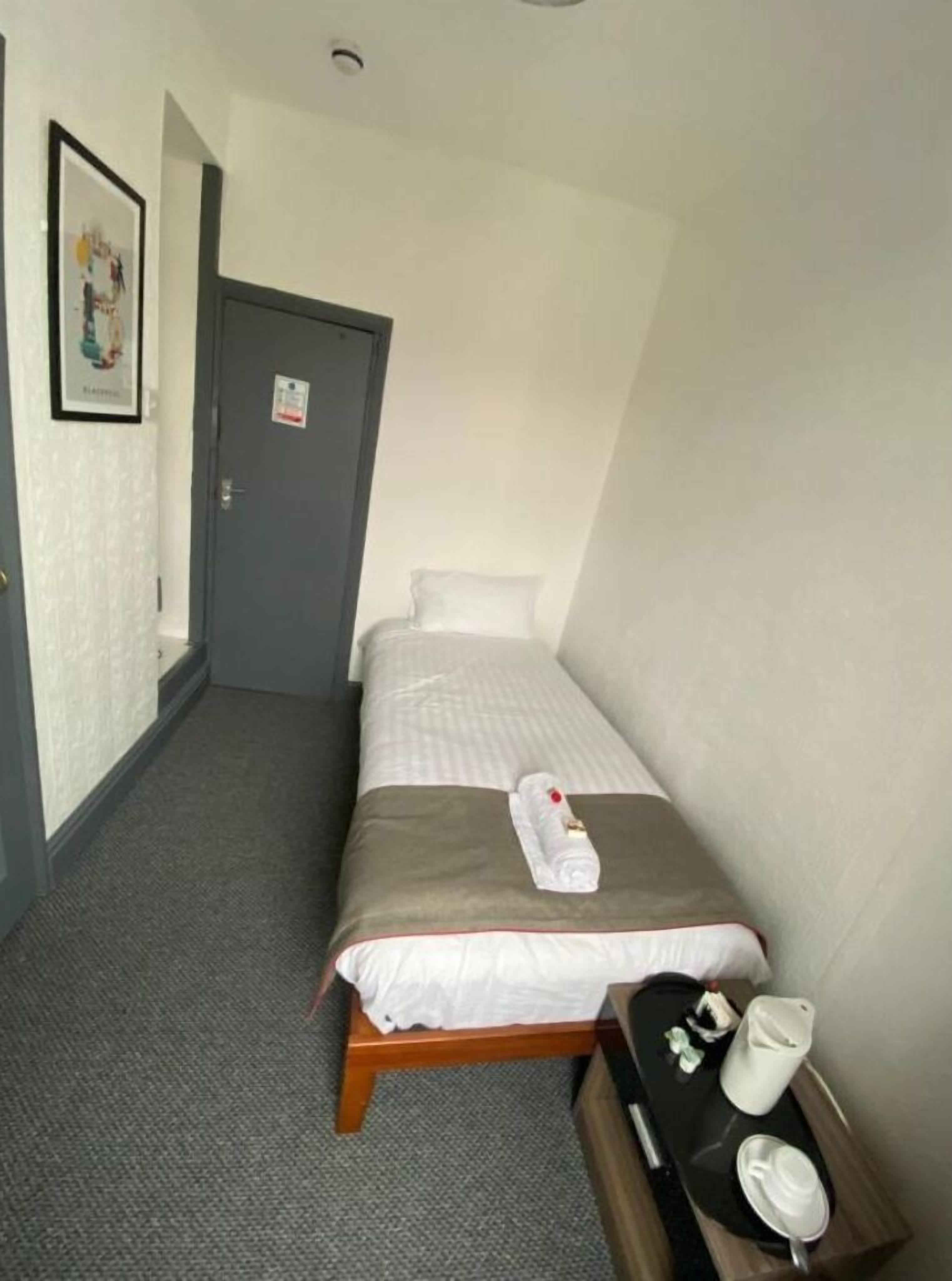A small, neatly arranged hotel room with a single bed, a bedside table with a tray containing a cup and a kettle, and a door with a sign.