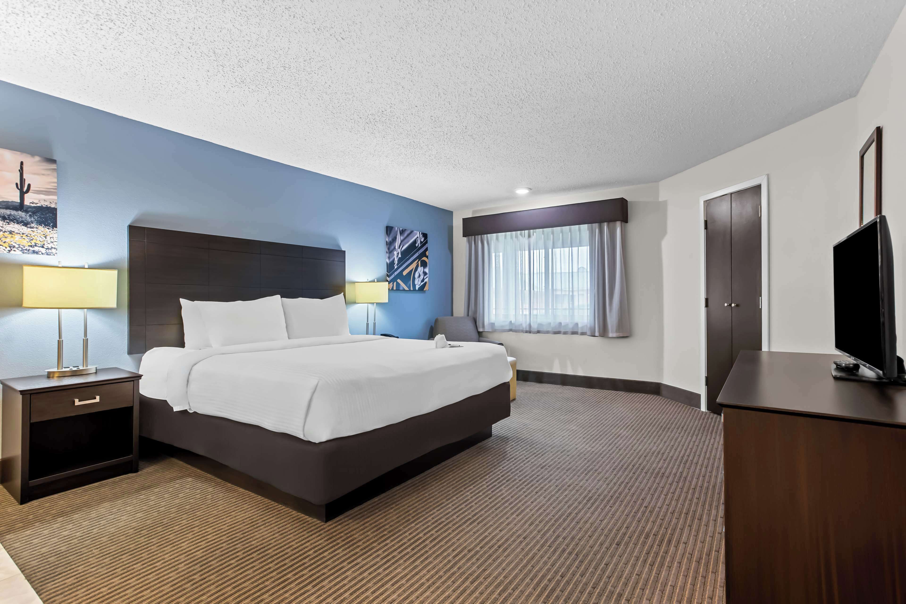 Habitación de hotel con una cama grande, una mesita de noche con lámpara, un televisor sobre un mueble de madera y una ventana con cortinas.