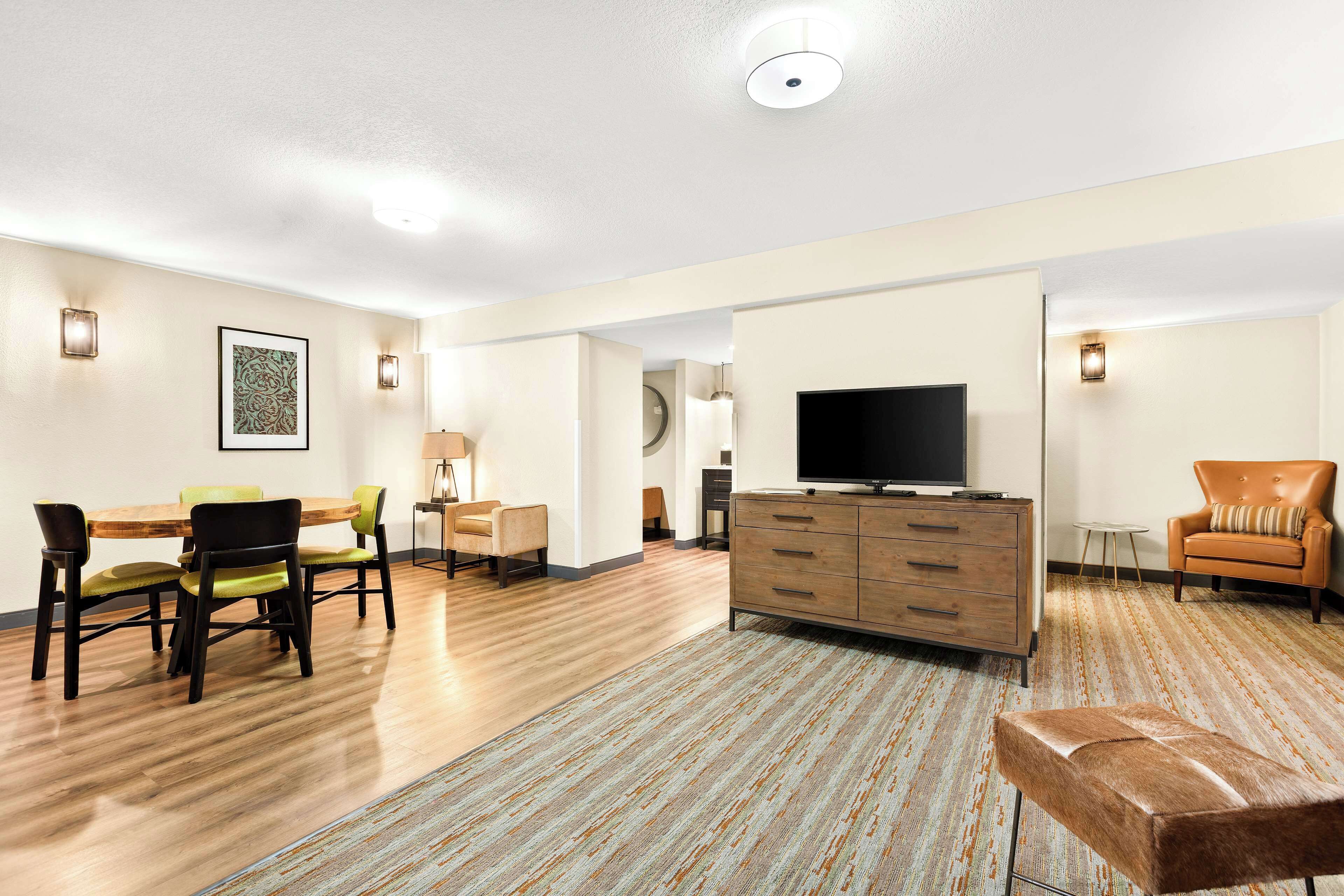 A modern living room with a wooden floor, a TV, a sofa, a dining table with chairs, and a patterned rug.