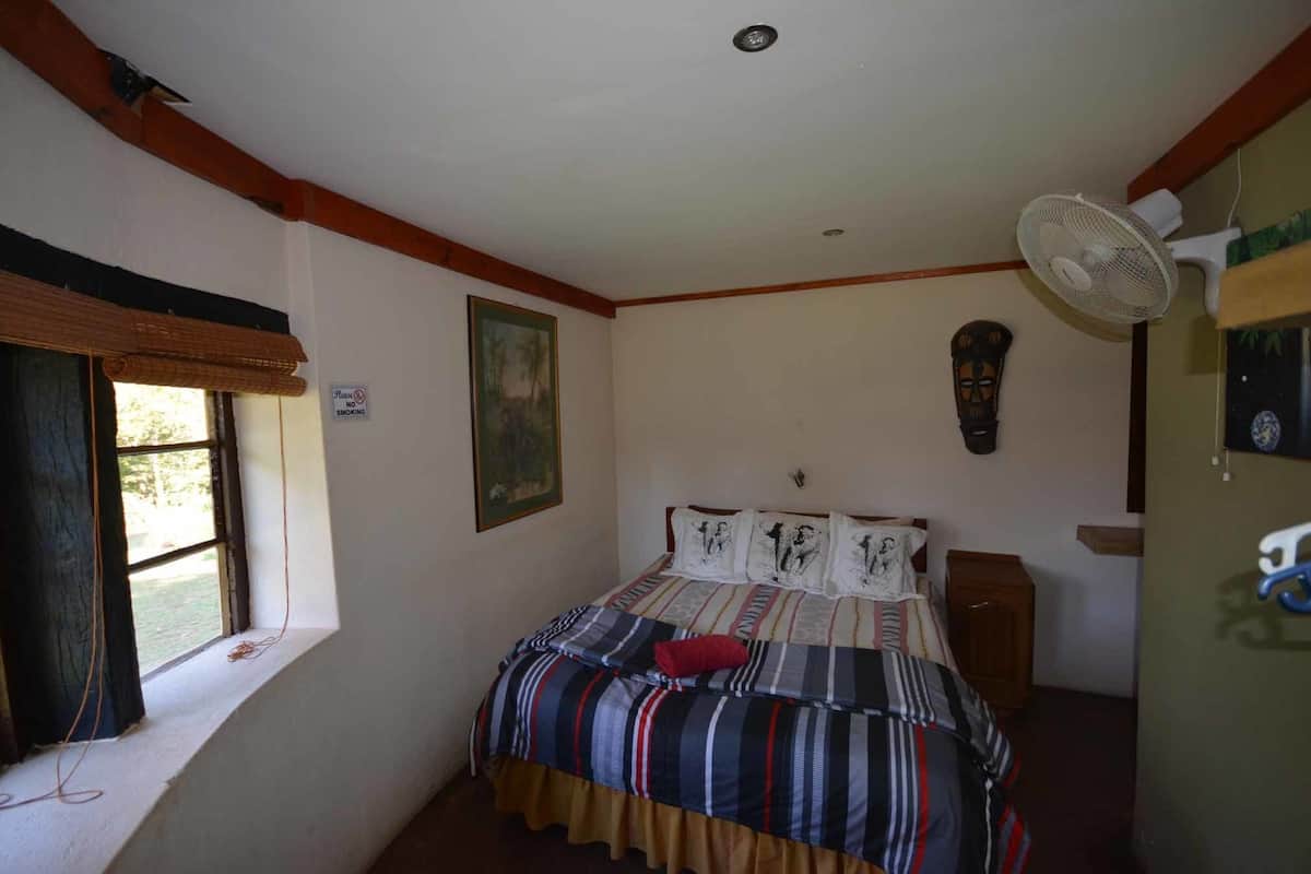 A bedroom with a bed, a window with a view, a fan, and a painting on the wall.