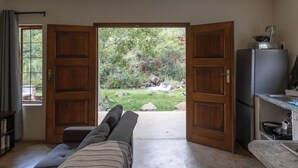 A room with a sofa facing a wooden door leading to a garden with a waterfall. - Rocky Drift Private Nature Reserve (Emgwenya)