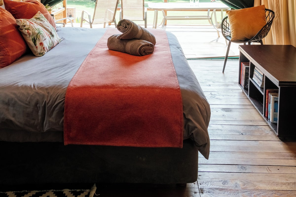 A tented accommodation with a bed, pillows, a rolled towel, a chair, and a table.