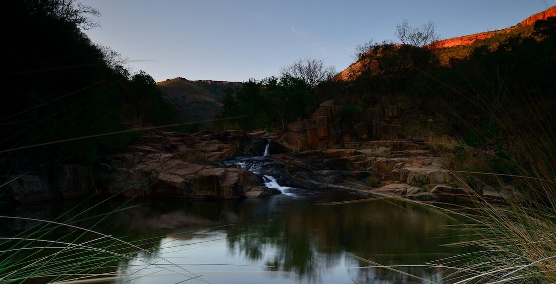 Paisaje natural con cascada, terreno rocoso y una masa de agua tranquila que refleja el entorno.