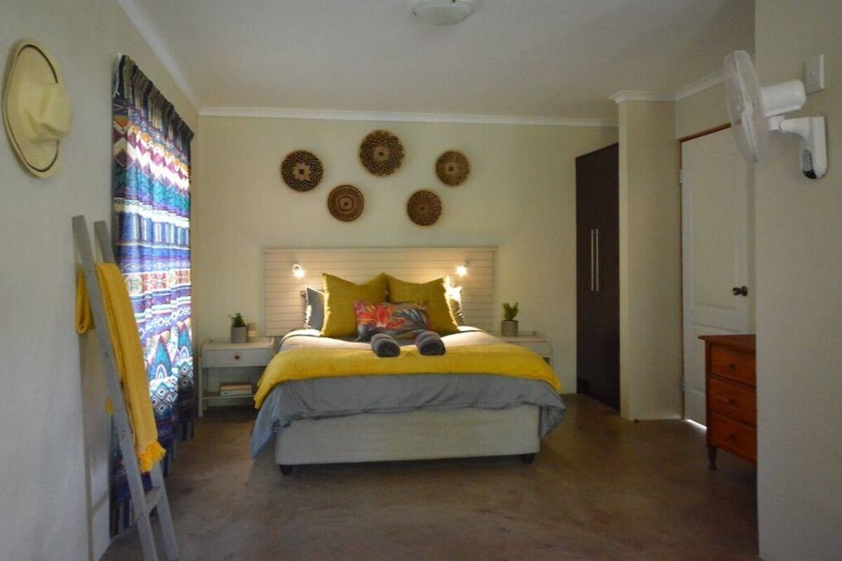 A bedroom with a bed, a nightstand, a dresser, a fan, and a door.