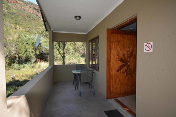 A balcony with a table and chairs, a door with a no-smoking sign, and a view of trees and hills. - Rocky Drift Private Nature Reserve (Emgwenya)