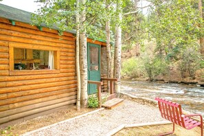 A log cabin with a window and door, a bench, and a river in the background. - Idlewilde by the River (Estes Park)