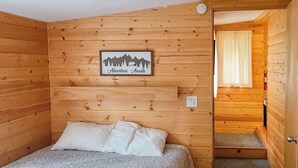 A wooden cabin room with a bed, a framed picture on the wall, and a door leading to another room. - Idlewilde by the River (Estes Park)