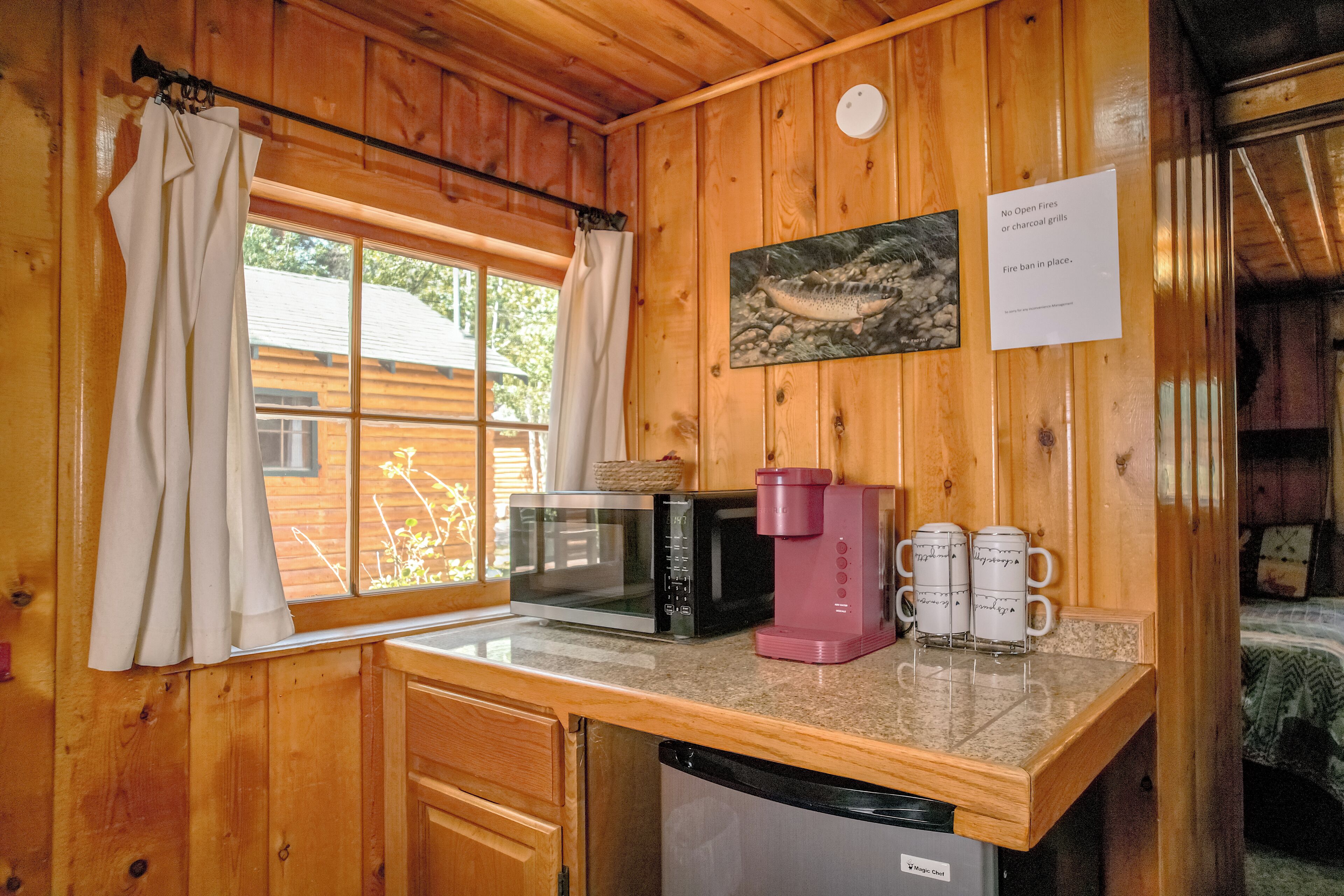 Una cocina compacta con paredes de madera, una ventana con cortinas, microondas, cafetera y lavavajillas.