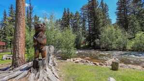 A wooden bear sculpture on a tree stump by a flowing river, surrounded by dense forest and a clear blue sky. - Idlewilde by the River (Estes Park)