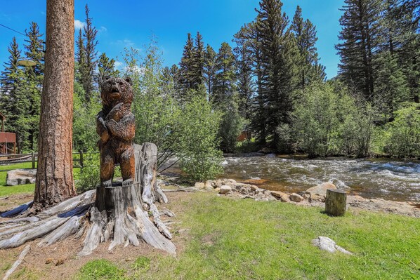 A wooden bear sculpture on a tree stump by a flowing river, surrounded by dense forest and a clear blue sky. - Idlewilde by the River (Estes Park)
