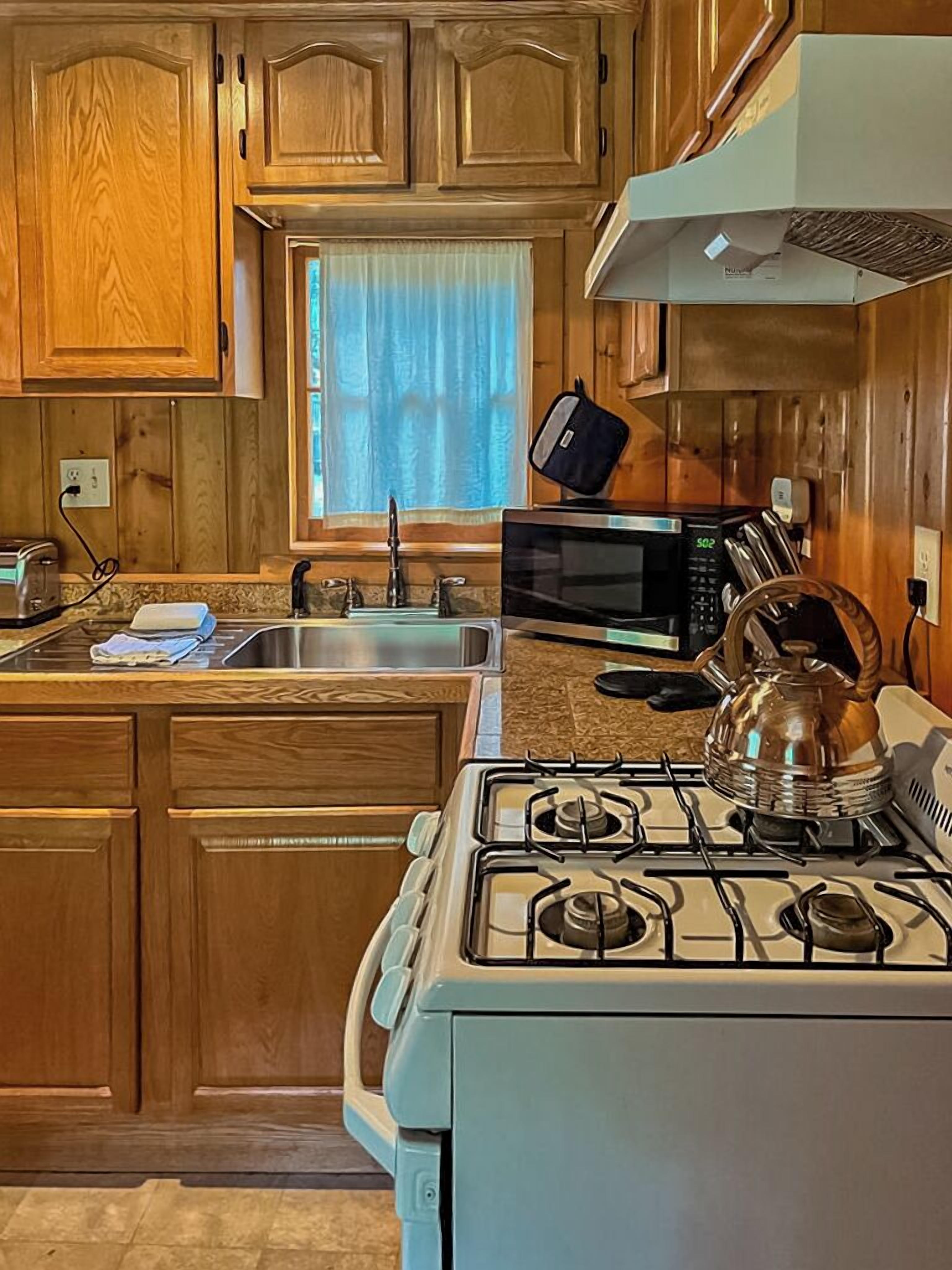 A kitchen with wooden cabinets, a gas stove, a microwave, and a window with curtains.